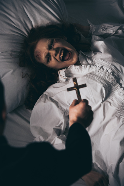 exorcist holding cross over creepy screaming demon in bed - Fotó, kép