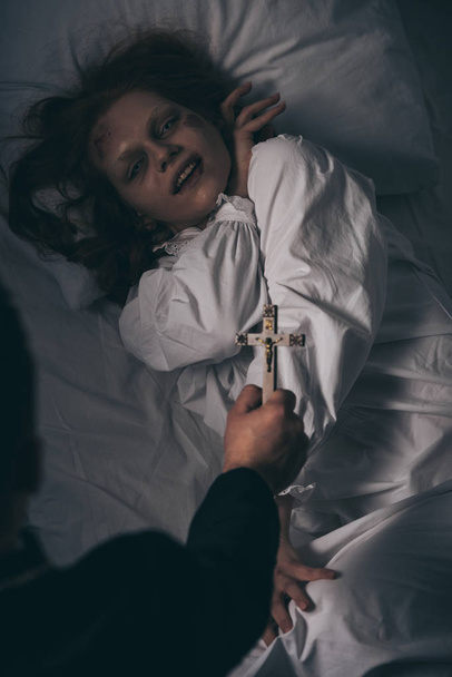 exorcist holding cross over demonic girl in bed - Photo, Image