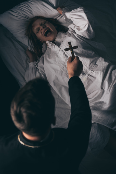ördögűző tartja kereszt felett megszállott kiabáló lány az ágyban - Fotó, kép