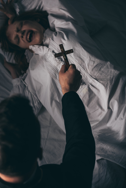 εξορκιστής κρατώντας σταυρό πάνω από εμμονή ουρλιάζοντας κορίτσι στο κρεβάτι - Φωτογραφία, εικόνα
