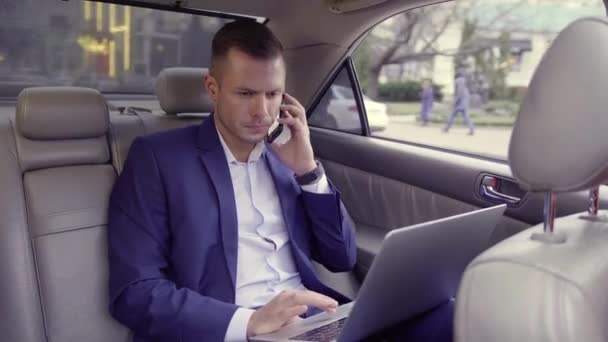 Portret of businessman in blue suit sitting in back seat of moving car, talking by phone phone while working on laptop while heading to office. Handsome beared male with strong face and blonde hair in - Materiał filmowy, wideo