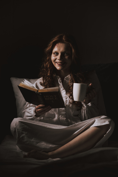 femme souriante démoniaque en chemise de nuit tenant bible et tasse sur le lit
 - Photo, image