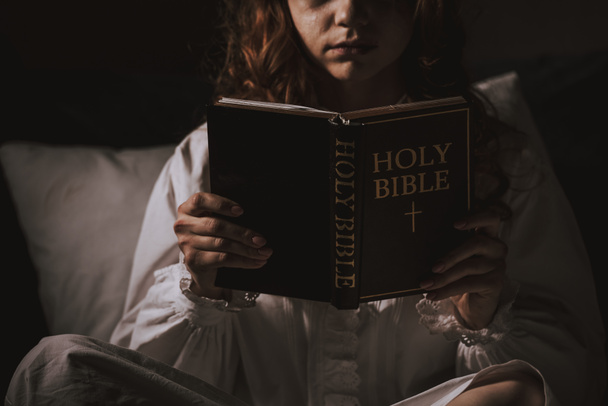 abgeschnittene Ansicht eines gruseligen Mädchens im Nachthemd, das Bibel auf dem Bett liest - Foto, Bild