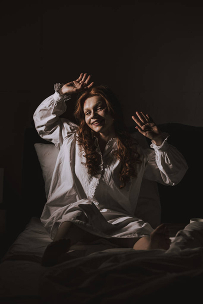 Τρομερή εμμονή χαμογελαστό κορίτσι σε νυχτικό κάθεται στο κρεβάτι - Φωτογραφία, εικόνα