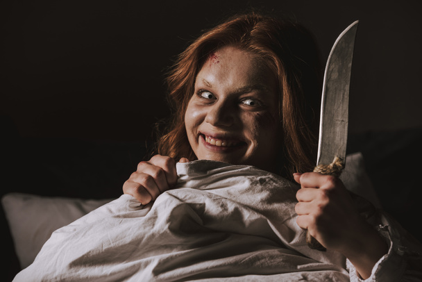 demoniacal smiling girl holding knife in bed - Photo, Image