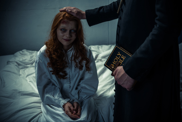 εξορκιστής κρατώντας τη Βίβλο και αγκαλιάζοντας δαιμονικό ανατριχιαστικό κορίτσι στην κρεβατοκάμαρα  - Φωτογραφία, εικόνα
