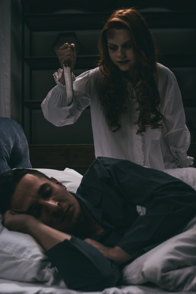παθιασμένη γυναίκα με μαχαίρι να στέκεται πάνω από έναν κοιμισμένο άντρα στην κρεβατοκάμαρα.  - Φωτογραφία, εικόνα