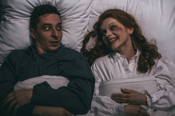 Top view of female smiling demon lying in bed with scared man
 - Фото, изображение