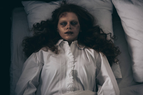 bovenaanzicht van geobsedeerd griezelig meisje in nachtjapon liggend in bed - Foto, afbeelding