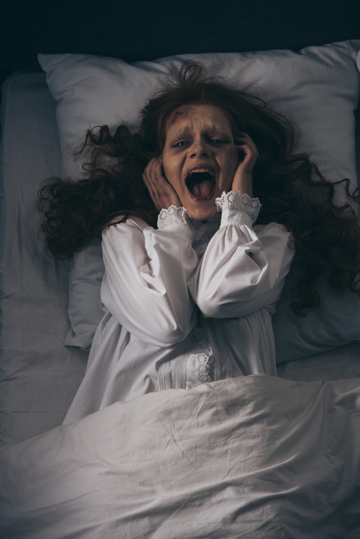 top view of obsessed girl in nightgown screaming while lying in bed - Foto, Imagen
