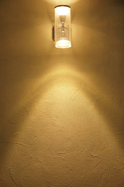 linterna en la pared amarilla se ilumina por la noche. Lámpara de poste contra una pared de estuco texturizada en la noche. escena noche calle
. - Foto, Imagen