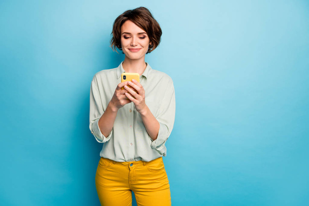 Photo of pretty lady holding telephone hands reading new blog post positive comments popular blogger wear casual green shirt yellow trousers isolated blue color background - Fotografie, Obrázek