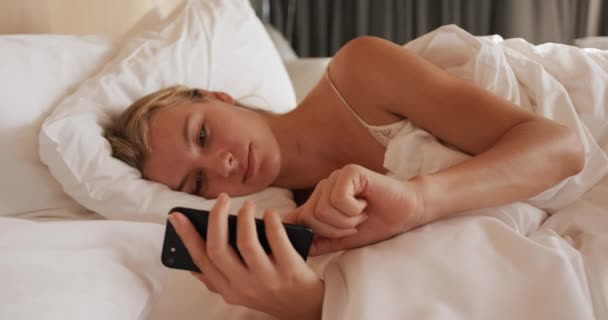 Front view of a Caucasian woman enjoying quality time in a hotel, lying on bed in bedroom and using smartphone, slow motion - Кадры, видео
