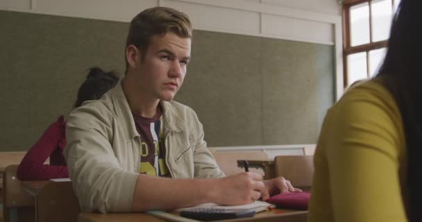 Side view of a teenage Caucasian boy in a school classroom sitting a desk, concentrating and raising his hand, with teenage classmates sitting at desks working in the background, in slow motion - Materiał filmowy, wideo