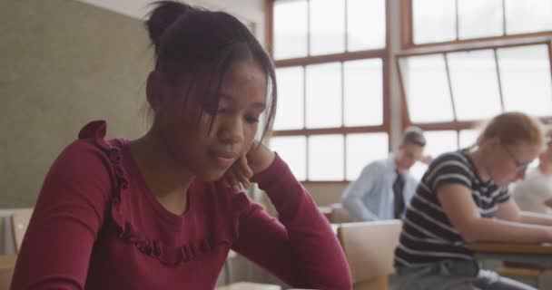 Side view close up of a teenage African American girl in a school classroom sitting a desk, smiling and concentrating, with a group of teenage male and female classmates sitting at desks working in the background, in slow motion - Filmagem, Vídeo