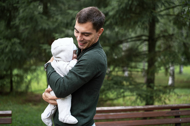 portret van lachende brunette vader in groene jas met baby in zijn armen in Park - Foto, afbeelding