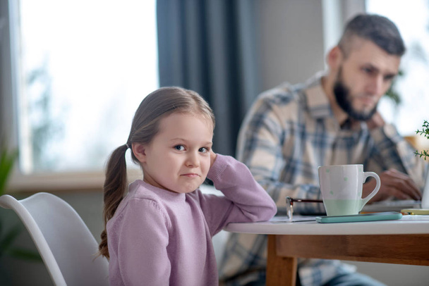 Little girl sitting at the table sadly smiling, dad busy. - Foto, Bild
