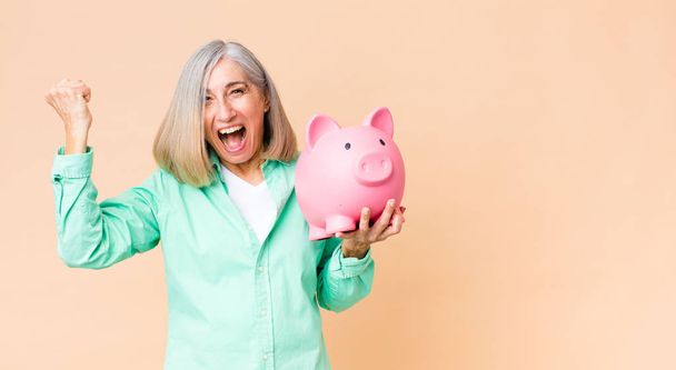 middle age pretty woman holding a piggy bank against copy space wall - Фото, изображение