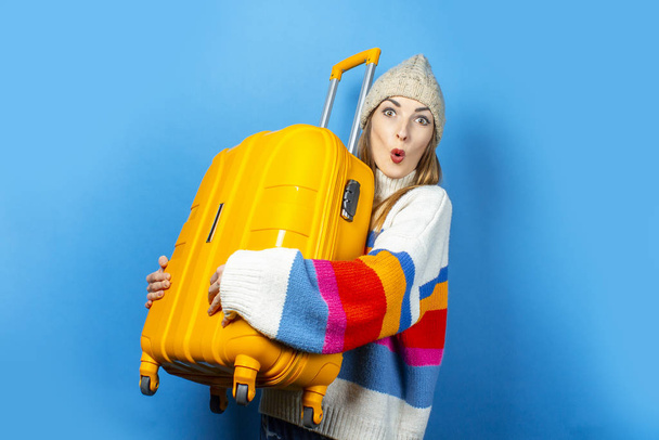 Şapkalı, şaşırmış yüzlü genç bir kadın mavi arka planda sarı bir bavul tutuyor. Seyahat, seyahat, tatil kavramı. Duygusallık. Pankart - Fotoğraf, Görsel