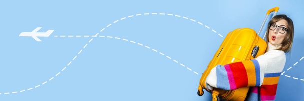 Egy meglepett arcú fiatal nő egy sárga bőröndöt tart kék alapon. Repülőgép ikon. Fogalom utazás, utazás, nyaralás. Érzelmileg. Zászlós - Fotó, kép