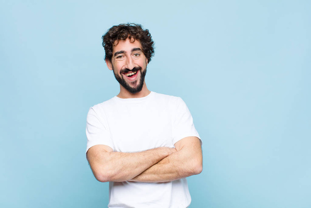 young crazy bearded man looking like a happy, proud and satisfied achiever smiling with arms crossed against flat wall - Foto, imagen