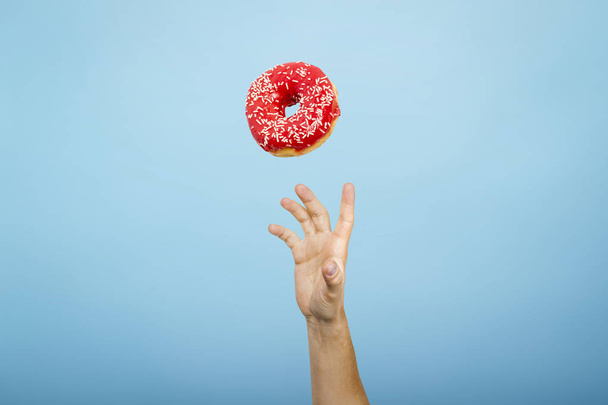 Hand fangen einen Donut mit rotem Zuckerguss. Blauer Papphintergrund. Das Konzept des Backens, handgemacht. Flache Lage, Draufsicht - Foto, Bild