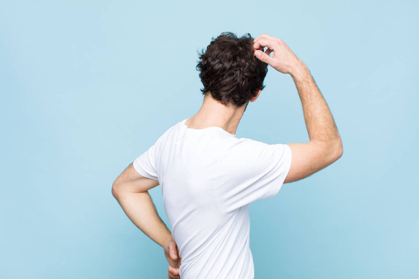 young crazy bearded man feeling clueless and confused, thinking a solution, with hand on hip and other on head, rear view against flat wall - Fotoğraf, Görsel