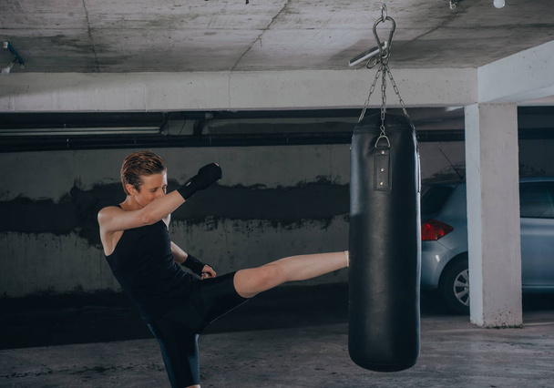 Caucasian woman in sportswear and with black bandages kicking the boxing bag inside a garage - Photo, Image