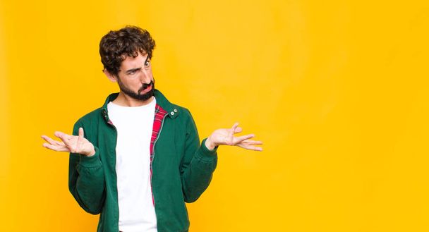 young crazy bearded man shrugging with a dumb, crazy, confused, puzzled expression, feeling annoyed and clueless against flat wall - Foto, Bild