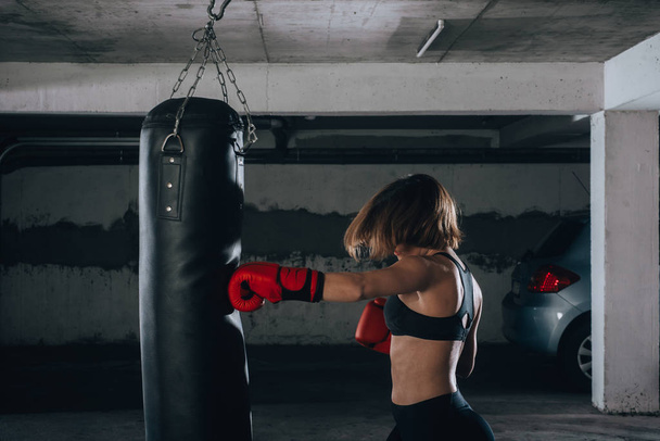 Vista del perfil de una joven fuerte golpeando una bolsa de boxeo dentro del garaje - Foto, Imagen