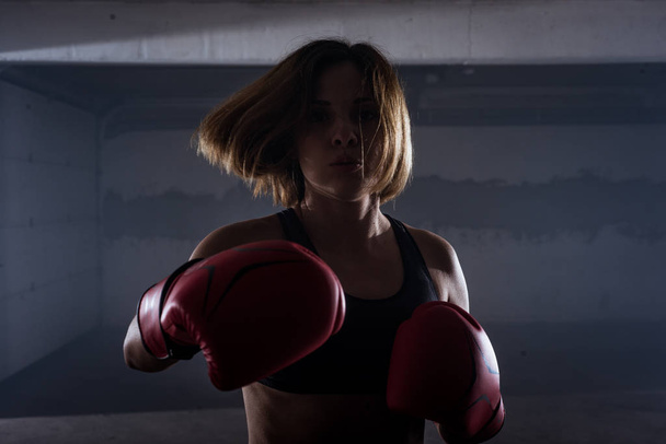 Силуэт портрет молодой привлекательной фитнес-женщины, занимающейся боксом в красных боксёрских перчатках в гараже - Фото, изображение