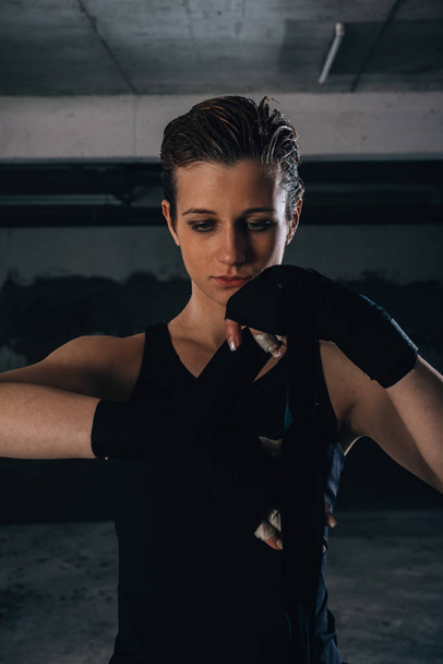 Retrato de una joven mujer fuerte enrollando vendajes negros en las muñecas, preparándose para el boxeo en un garaje. - Foto, Imagen