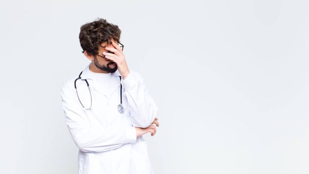 young doctor man looking stressed, ashamed or upset, with a headache, covering face with hand against copy space wall - Foto, imagen
