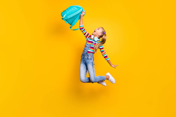 Full size photo crazy energetic kid jump hold blue bag rucksack enjoy primary school lessons wear striped sweater jumper denim jeans sneakers isolated bright shine yellow color background - Foto, Bild