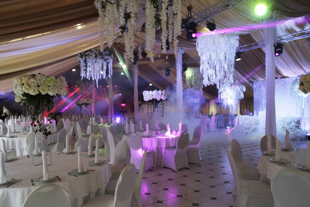 een zaal voor een banket in het wit, versierd met bloemstukken, verlicht door de stralen van discolampen - Foto, afbeelding