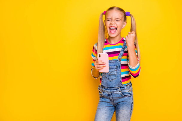 Portrait of delighted kid use cellphone win social network lottery raise fists scream yeah wear striped sweater jumper denim jeans isolated over shine yellow color background - Foto, Imagem