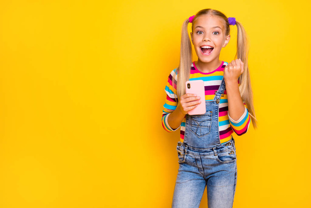 Surprised crazy energetic preteen kid use smartphone read social media news win lottery raise fists scream yes wear striped sweater jumper denim jeans isolated yellow bright color background - Zdjęcie, obraz