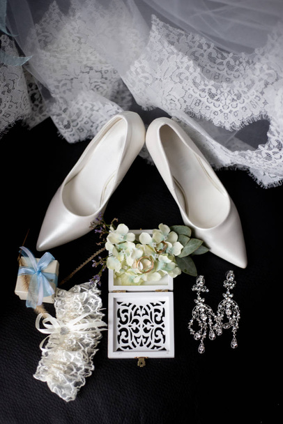 bruiloft accessoires: trouwringen in een decoratieve doos met bloemen, oorbellen, damesschoenen, een kousenband van de bruid, een klein geschenk, kant van een trouwjurk - Foto, afbeelding