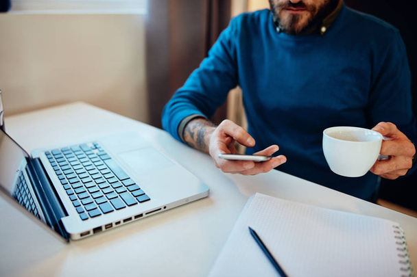 Mladý pohledný vousatý hipster sedí ve své kanceláři, drží šálek kávy a používá chytrý telefon. - Fotografie, Obrázek