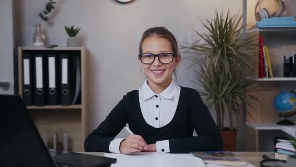Front view of smiling happy likable schoolgirl in uniform which sitting over her homework and thinking about a idea while looking at camera - Кадри, відео