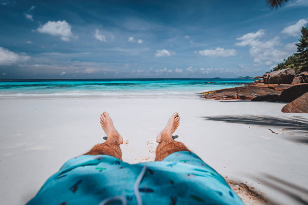 POV of male wearing swimming shorts with tanned legs on paradise white sand tropical exotic beach with view to turquoise blue ocean. Travel holidays vacation concept - Фото, зображення