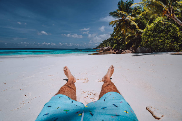 Male wearing swimming shorts with tanned legs on paradise white sand tropical exotic beach with view to turquoise blue ocean. Travel holidays vacation concept - Фото, изображение
