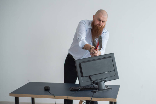 Злой лысый мужчина с рыжей бородой в офисе в деловом костюме разбивает топор компьютером. Менеджер, сконцентрированный на нервном срыве, ломает монитор
. - Фото, изображение