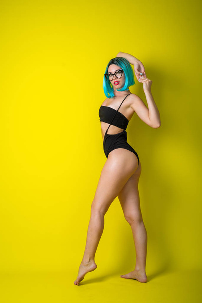 Beautiful woman in a black bikini in a colored wig and sunglasses on a yellow background. Girl posing in the studio. - Fotó, kép