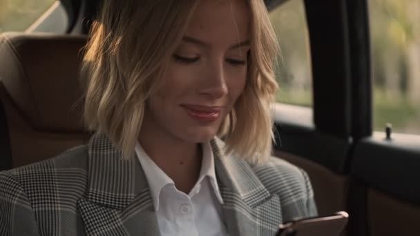 Close up view of Cheerful blonde business woman in coat using smartphone while sitting in car - Footage, Video