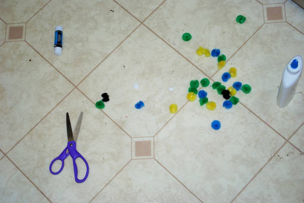 Multi colored cotton pompoms, glue, glue sticks, construction paper, and scissors covering a linoleum floor during arts and crafts time - Photo, Image