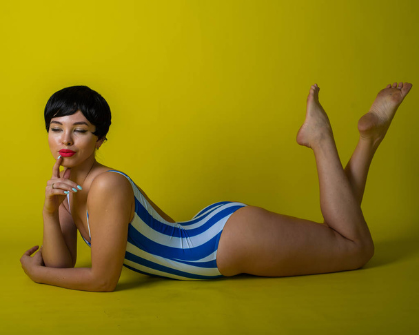 Attractive brunette lies on her stomach in a striped bikini. Short-haired young woman in bodysuit on a yellow background in the studio. - Foto, imagen