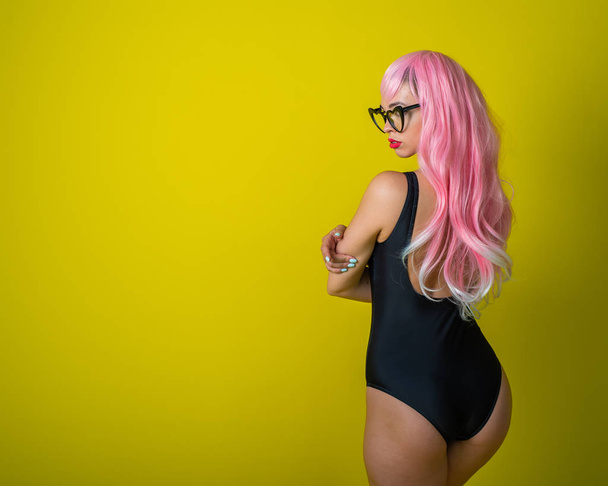 A woman with long wavy artificial hair is standing back in a black swimsuit. Fashion girl in a curly pink wig on a yellow background. - Valokuva, kuva