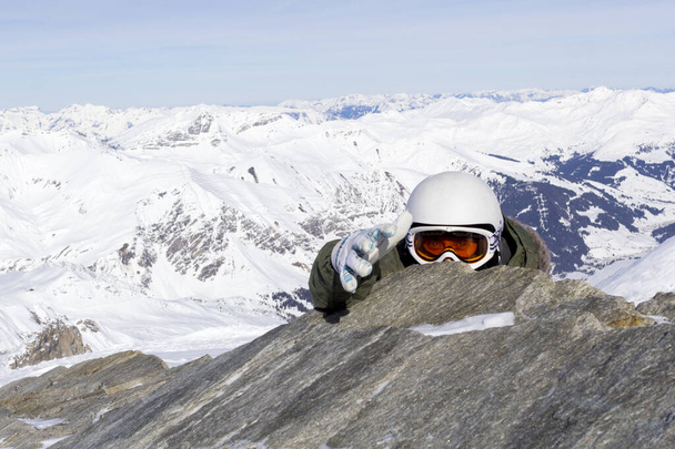 Snowboarder σε πορτοκαλί γυαλιά σκι σε ένα λευκό κράνος ανεβαίνει στην κορυφή και επεκτείνει ένα χέρι βοήθειας στις Άλπεις βουνά. Στο φόντο των βουνών. - Φωτογραφία, εικόνα