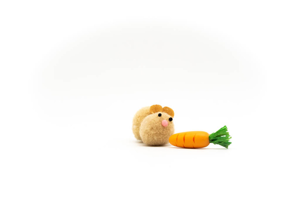 Roztomilý hnědý pom pom králík hračka při pohledu toužebně na oranžovou mrkev - Fotografie, Obrázek